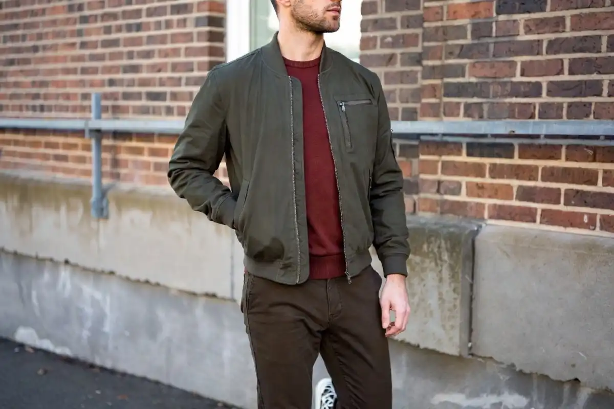 Man in olive bomber jacket layered over burgundy sweater and dark chinos in an urban fall street setting